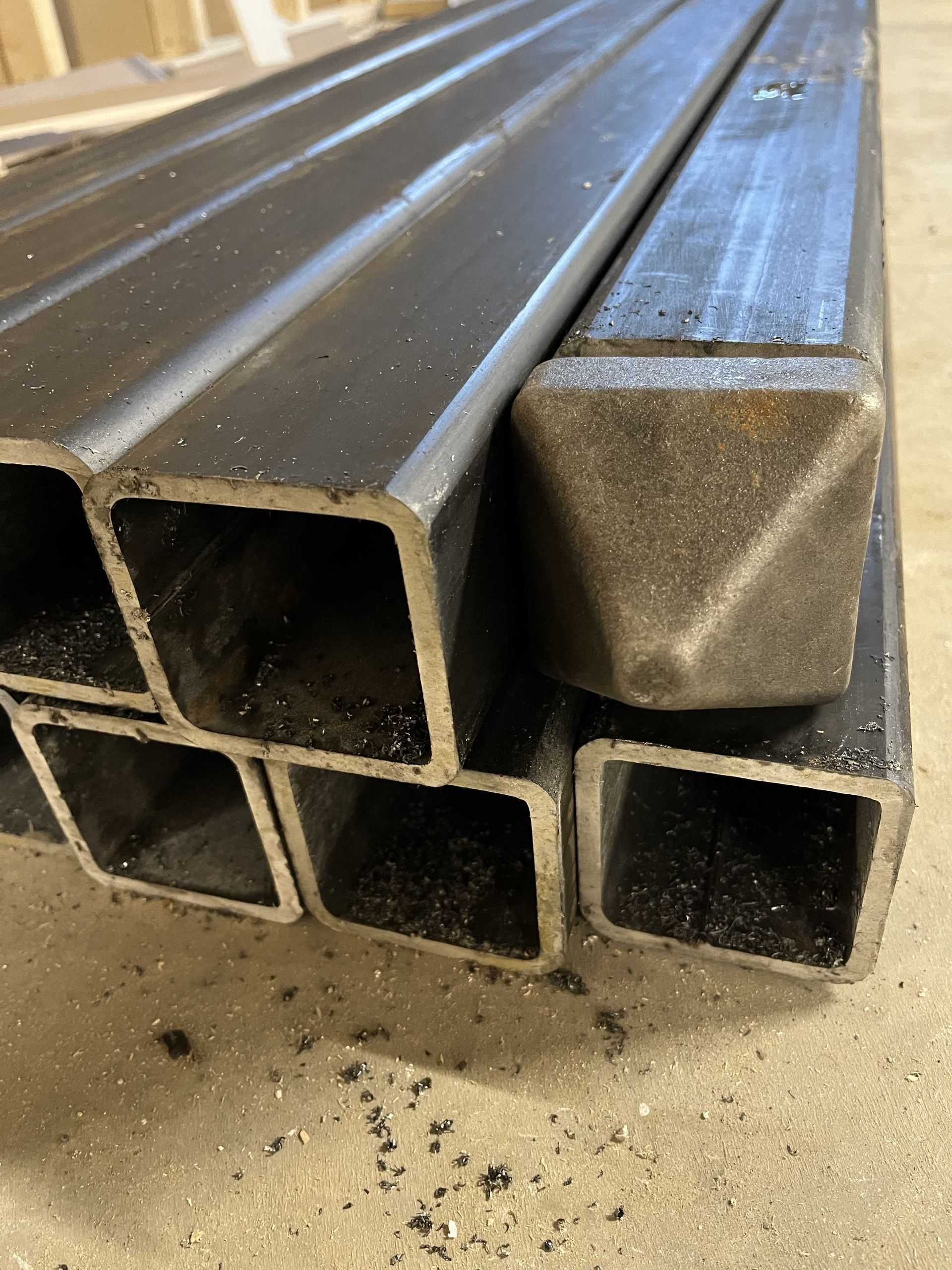 Un empilement de tubes carrés en acier sur un sol en béton, l'un des tubes étant coiffé d'un raccord métallique en forme de pyramide.