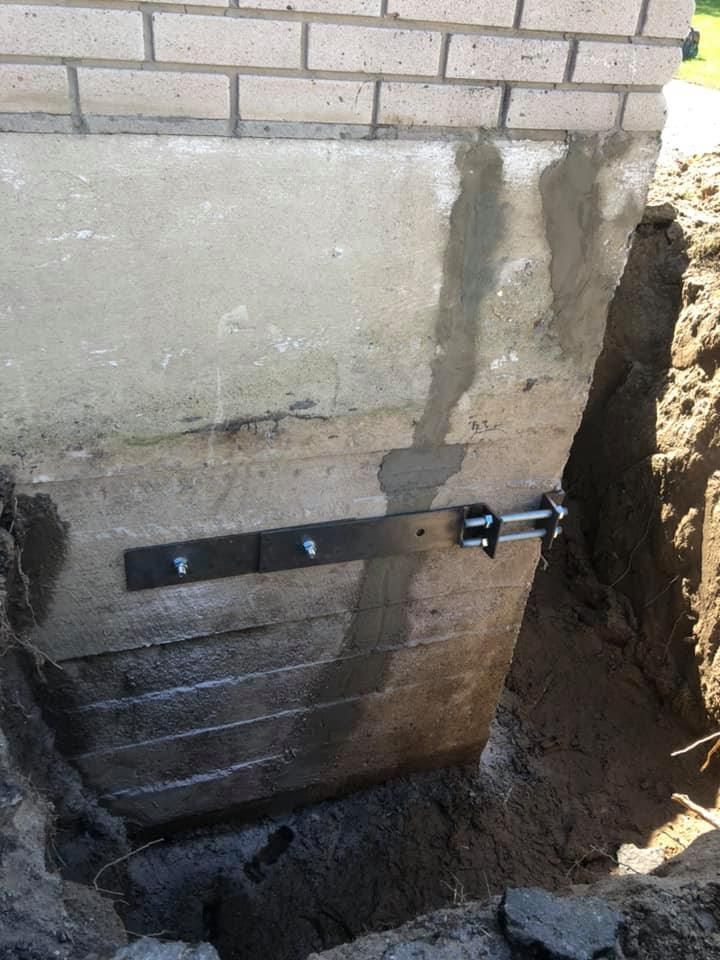 Un étai métallique de réparation murale ancré dans un mur de fondation en béton, avec une profonde excavation visible sur la droite.