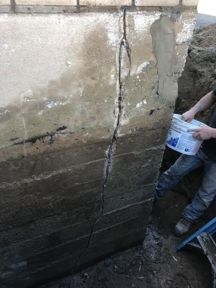 Une personne tient un seau tout en se tenant à côté d'un mur de fondation présentant une grande fissure structurelle verticale.