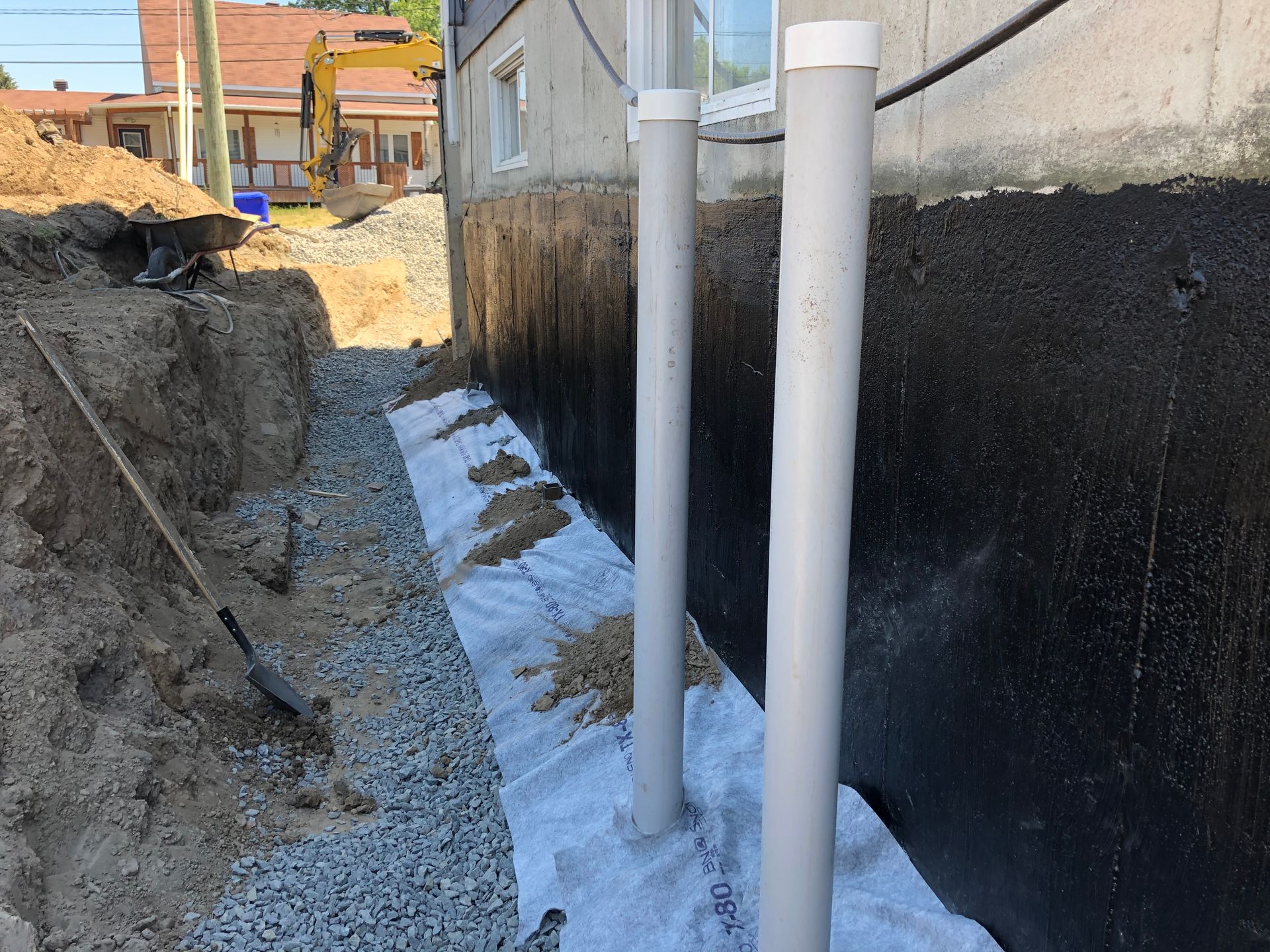 Une tranchée le long d'un mur de fondation avec un revêtement imperméable, des tuyaux de drainage et une couche de géotextile filtrante sur du gravier.
