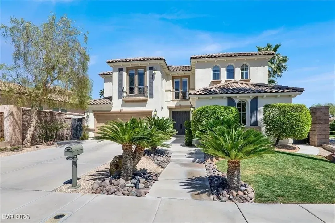 Two-story tan stucco house with a palm-lined driveway and desert landscaping