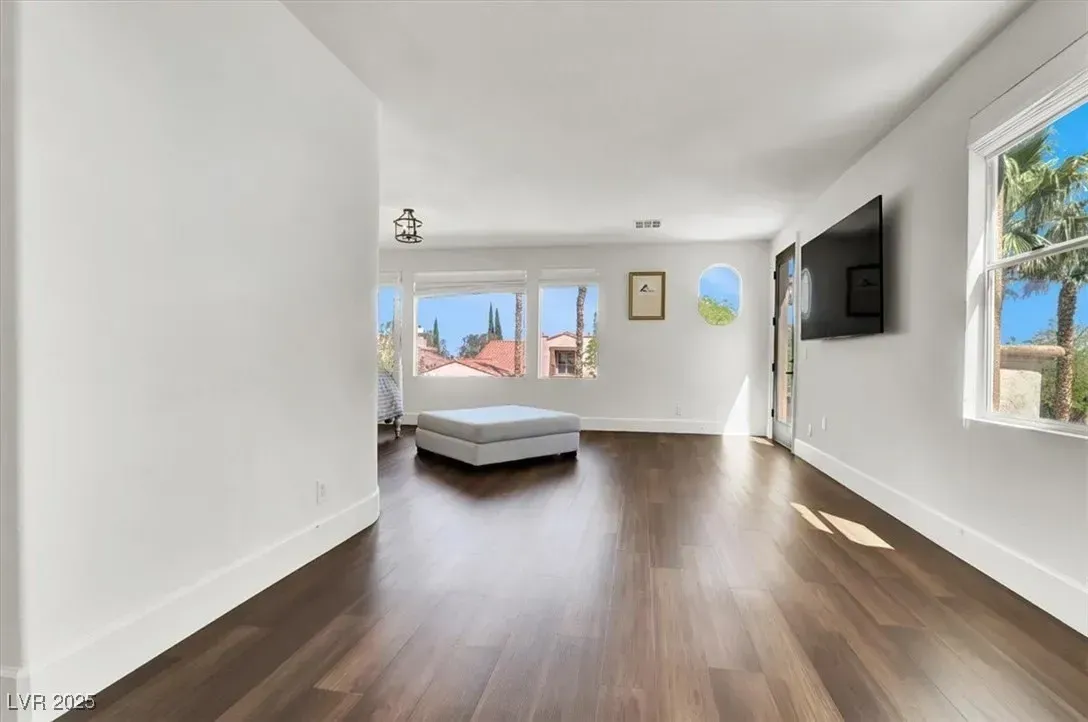 Bright empty bedroom with dark wood floors, a bed by windows, and a wall-mounted TV.