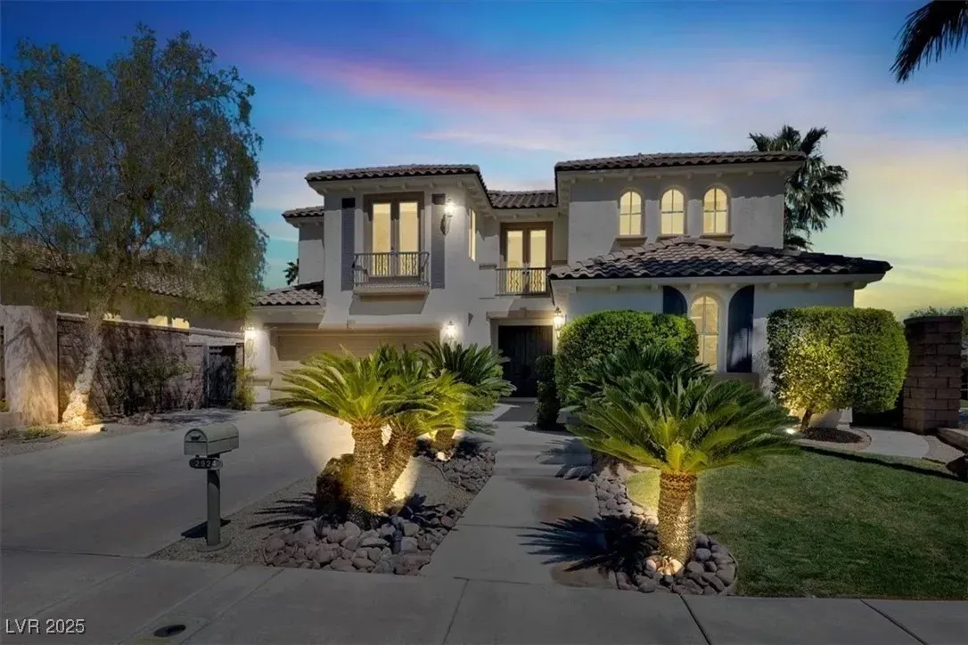 Two-story stucco house at dusk with lit windows, palm trees, and a landscaped driveway