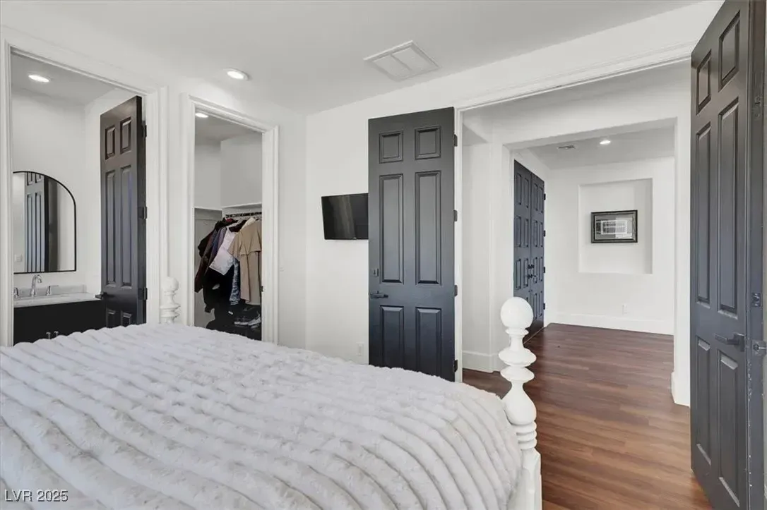 Bright bedroom with white bedding, dark doors, mirrored closet, and hardwood floors leading to a hallway