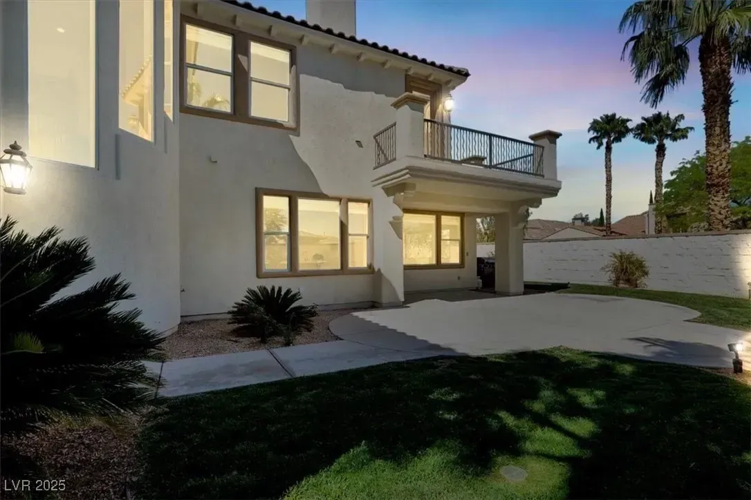 Modern two-story stucco house at dusk with lit windows, balcony, driveway, and palm trees.