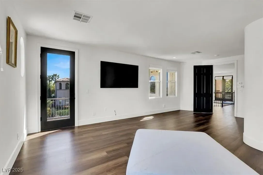 Bright empty bedroom with wall-mounted TV, hardwood floors, white walls, and glass doors to a balcony
