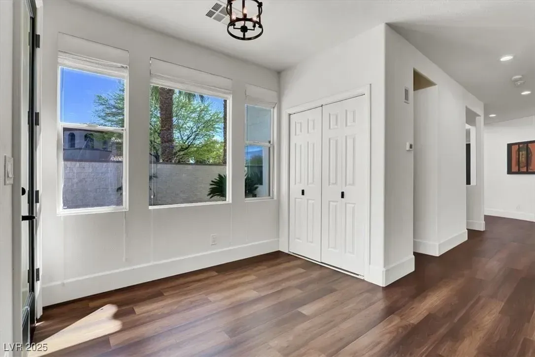 Bright empty room with white walls, dark wood floors, windows, closet, and hallway entrance