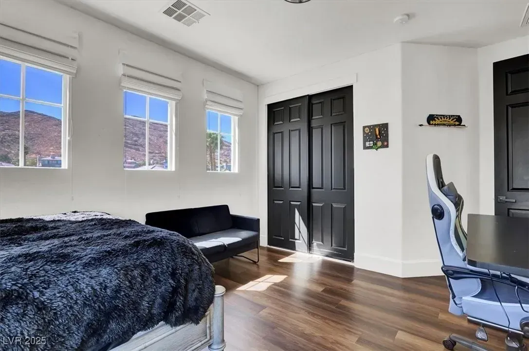 Bright bedroom with white walls, wood floors, black bed and couch, and sunlight through three windows.
