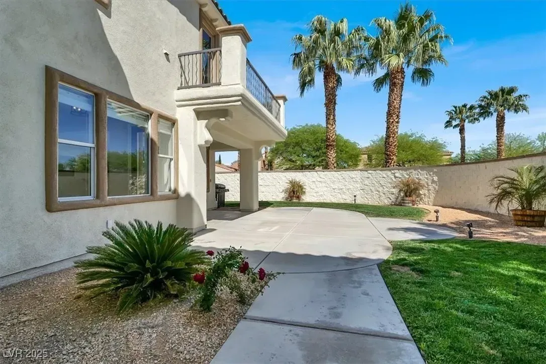 Sunny backyard patio beside a stucco house with palm trees, grass, and a stone wall
