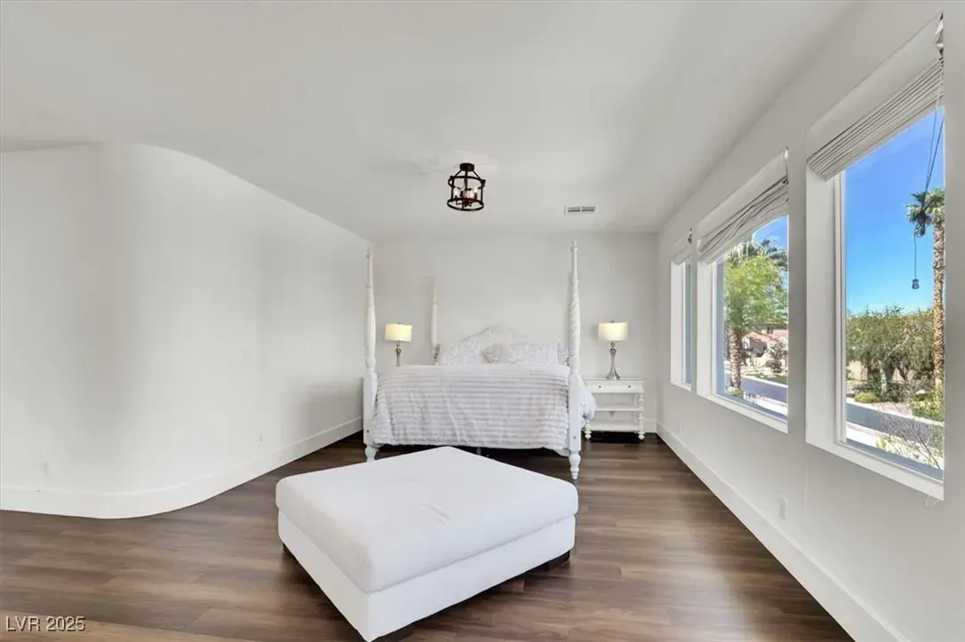 Bright white bedroom with a bed, large windows, dark wood floor, and a white ottoman at the foot of the bed
