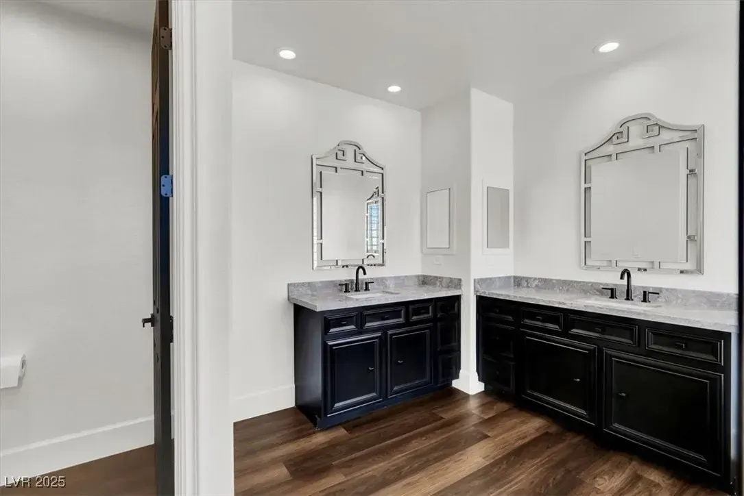 Modern bathroom with two black vanities, ornate mirrors, white walls, and wood flooring.