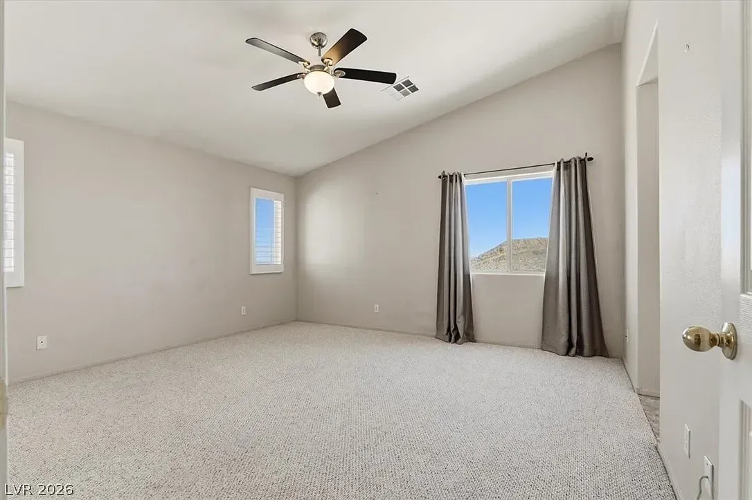 Empty beige bedroom with carpet, ceiling fan, and window with gray curtains