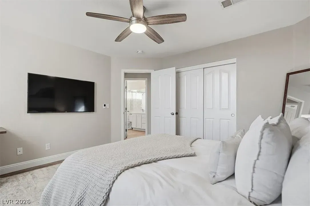 Bright bedroom with a wall-mounted TV, ceiling fan, white bedding, and open door to a bathroom and closet.