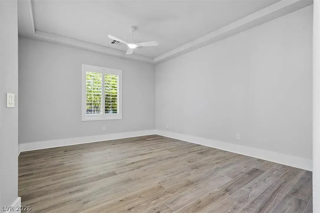 Empty light gray bedroom with wood flooring, white trim, and a window under a ceiling fan.