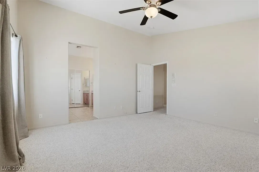 Empty carpeted bedroom with white walls, ceiling fan, and open doorways to a bathroom and closet