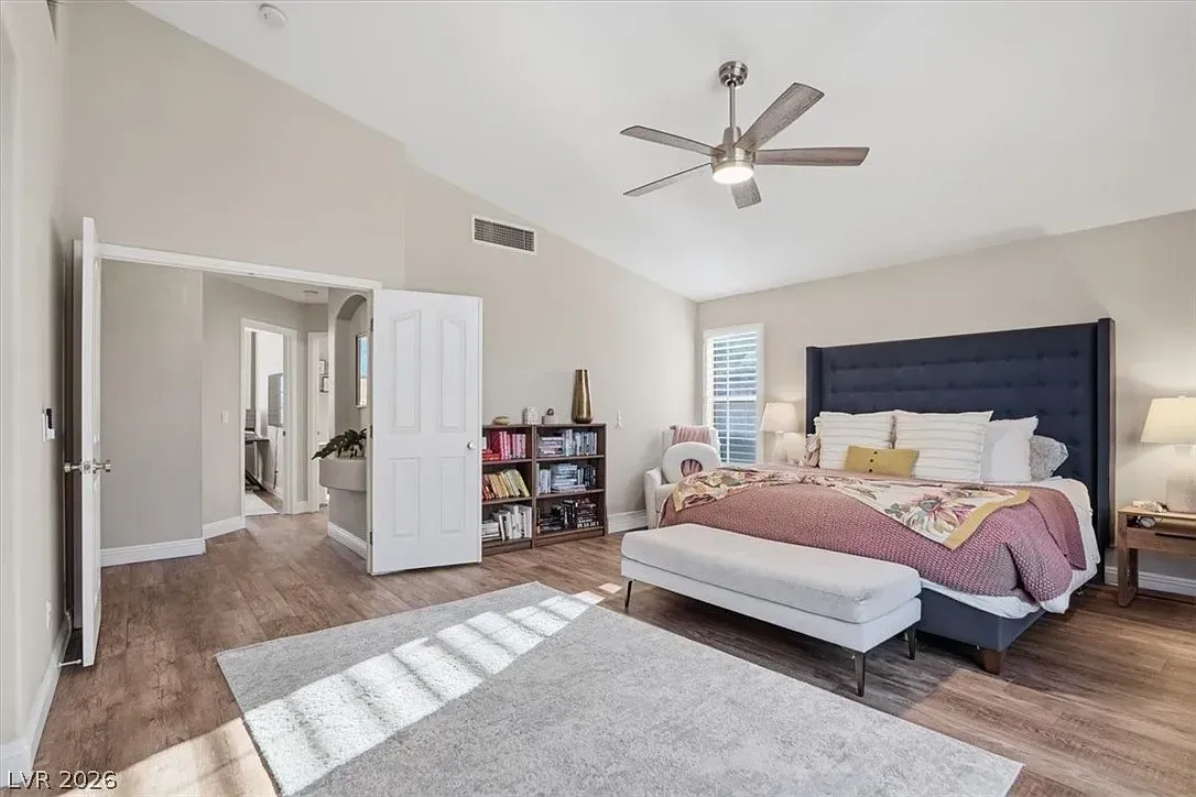 Spacious bedroom with navy upholstered bed, white dresser, ceiling fan, and sunlight on rug and wood floor