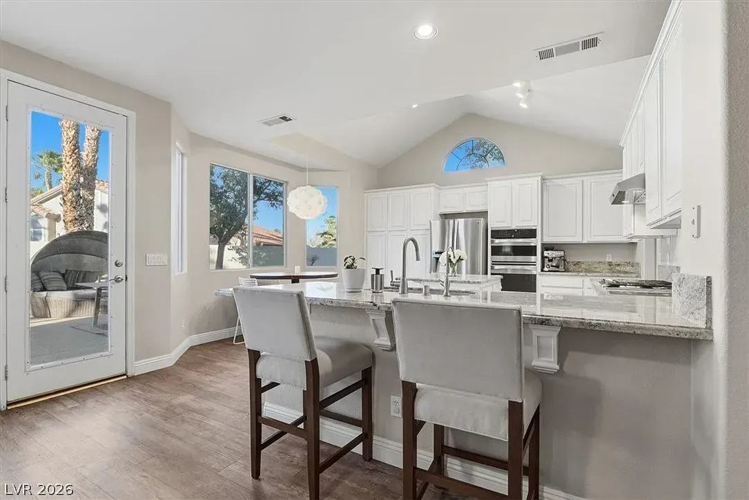 Bright modern kitchen with white cabinets, granite island, and two gray barstools by a sunlit door.