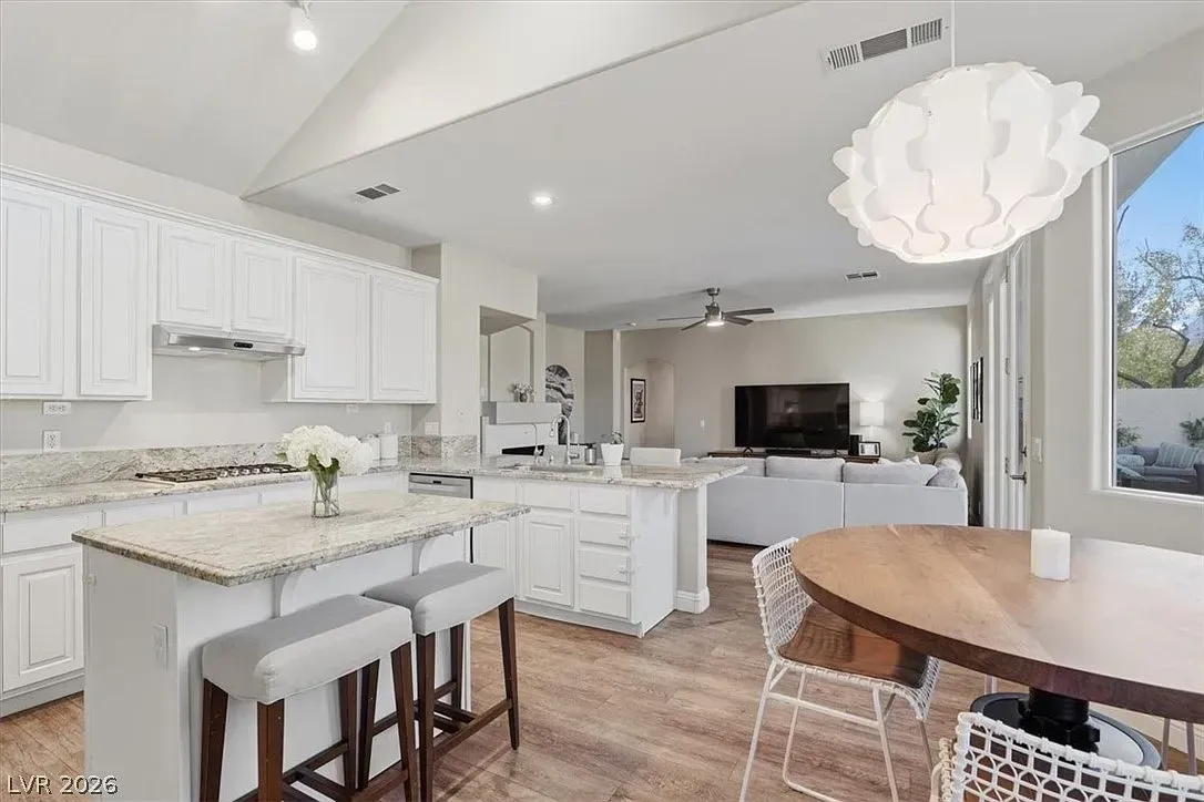 Bright white kitchen with granite island, bar stools, and dining area beside a window