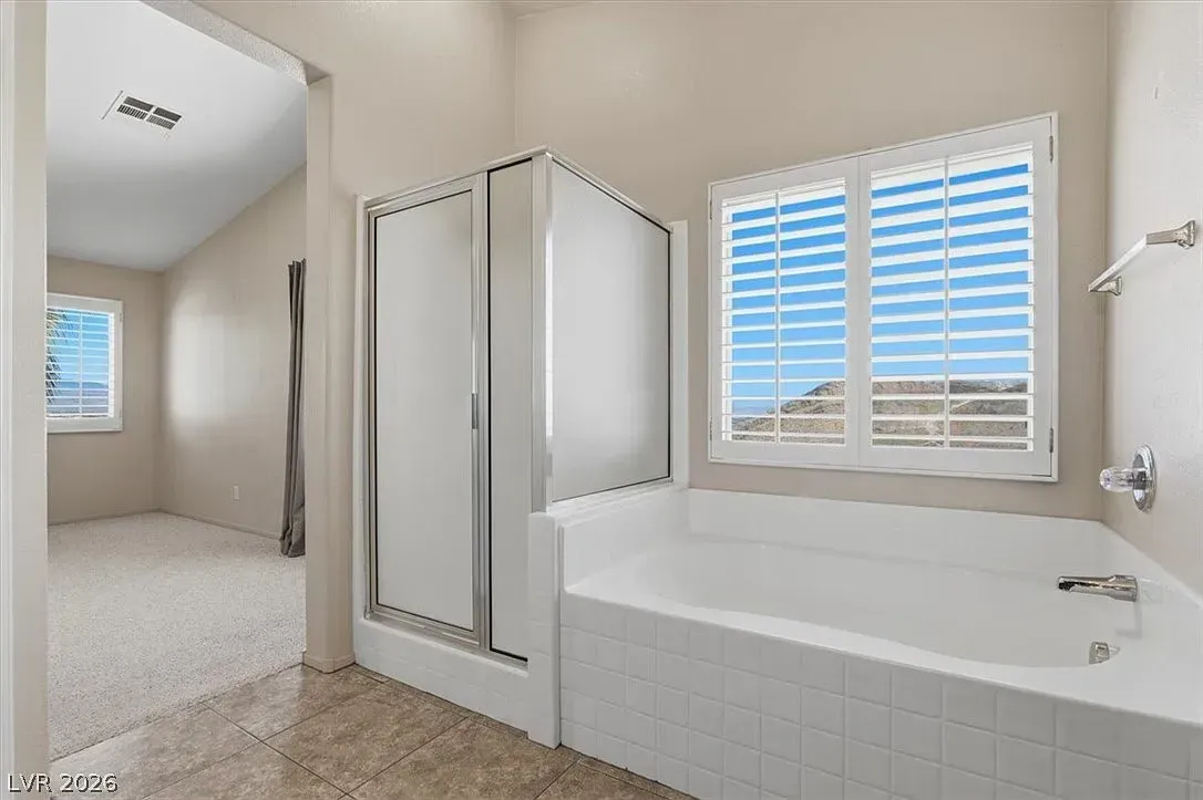 Bright bathroom with a glass shower, white tub, window shutters, and beige tile floor.