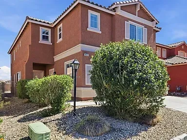Two-story tan stucco house with white trim, front shrubs, and a rock landscaped yard