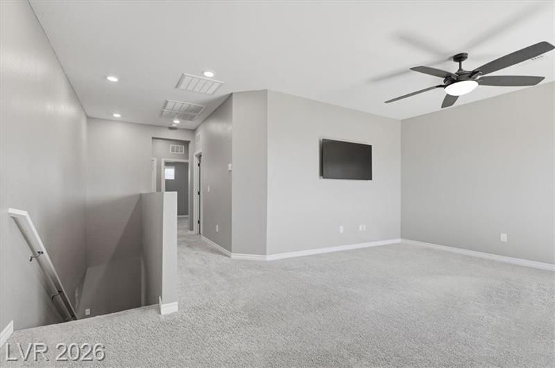 Empty gray carpeted upstairs room with ceiling fan, wall-mounted TV, and stairwell railing