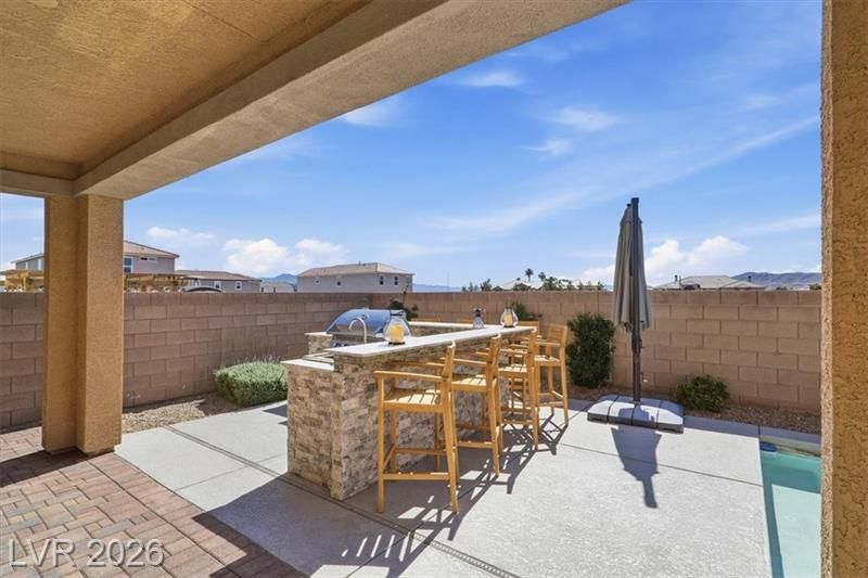 Covered patio with a stone outdoor bar, stools, and a closed umbrella beside a pool under a blue sky.