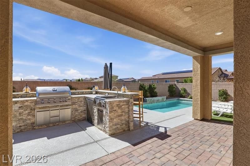 Covered patio with built-in grill, stone counter, and pool view in a sunny backyard