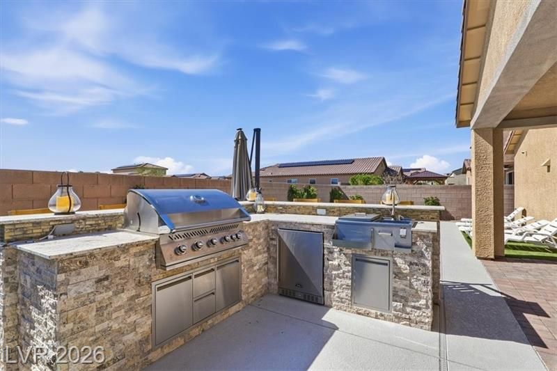 Outdoor stone patio with built-in stainless steel grill and sink beside a house, under a blue sky.