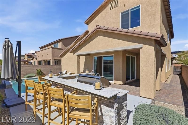 Backyard patio with outdoor dining table, bar stools, grill, and a covered seating area beside a tan house