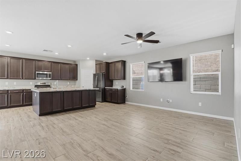 Open-plan kitchen and living room with dark cabinets, light wood floors, ceiling fan, and wall-mounted TV