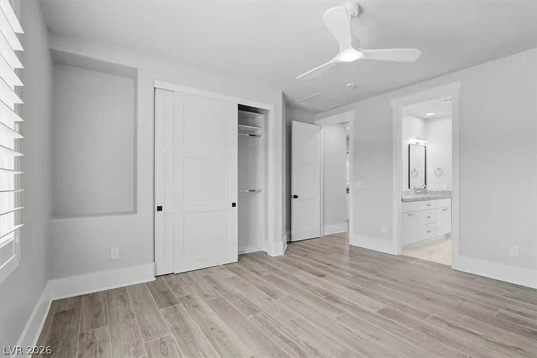 Bright empty bedroom with wood flooring, white walls, ceiling fan, and open closet leading to a bathroom.