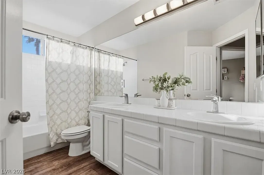 Bright white bathroom with double vanity, large mirror, shower curtain, and toilet by a window
