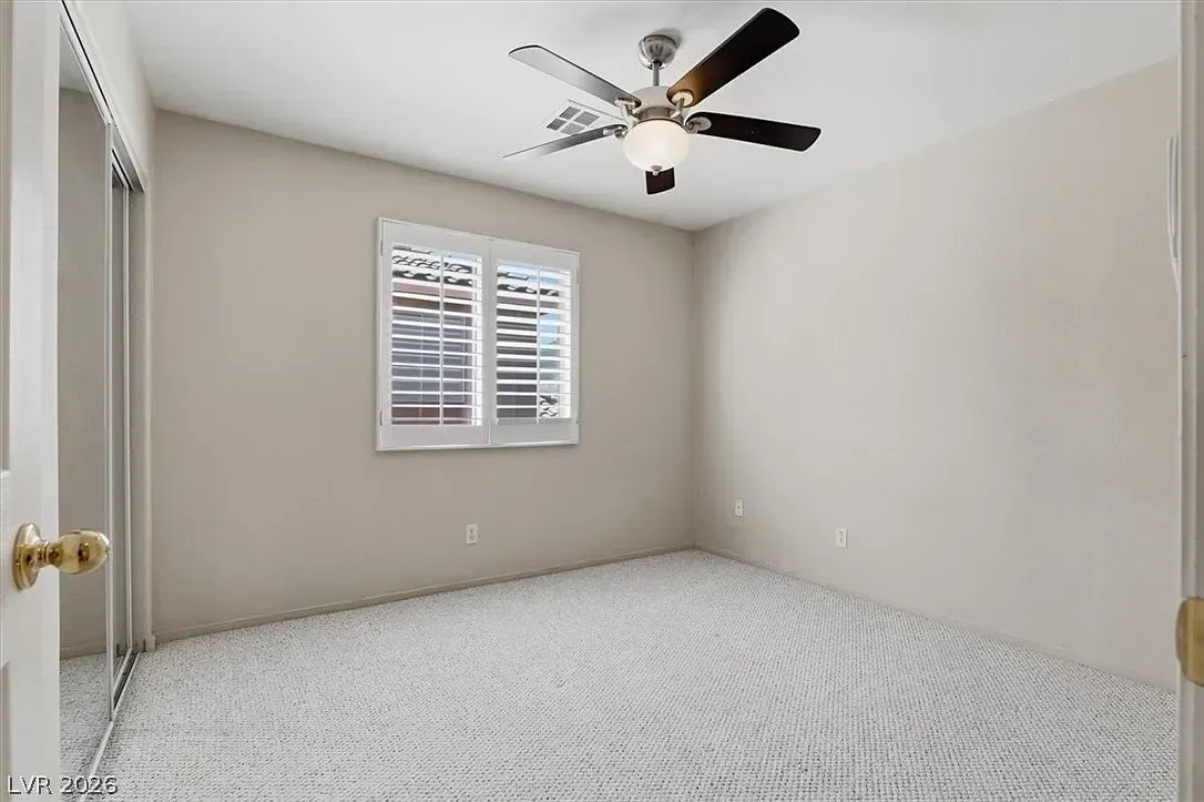 Empty beige bedroom with carpet, ceiling fan, and window with white shutters