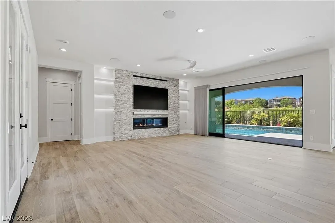 Spacious modern living room with stone fireplace, light wood floors, and open glass doors to a pool and lake view