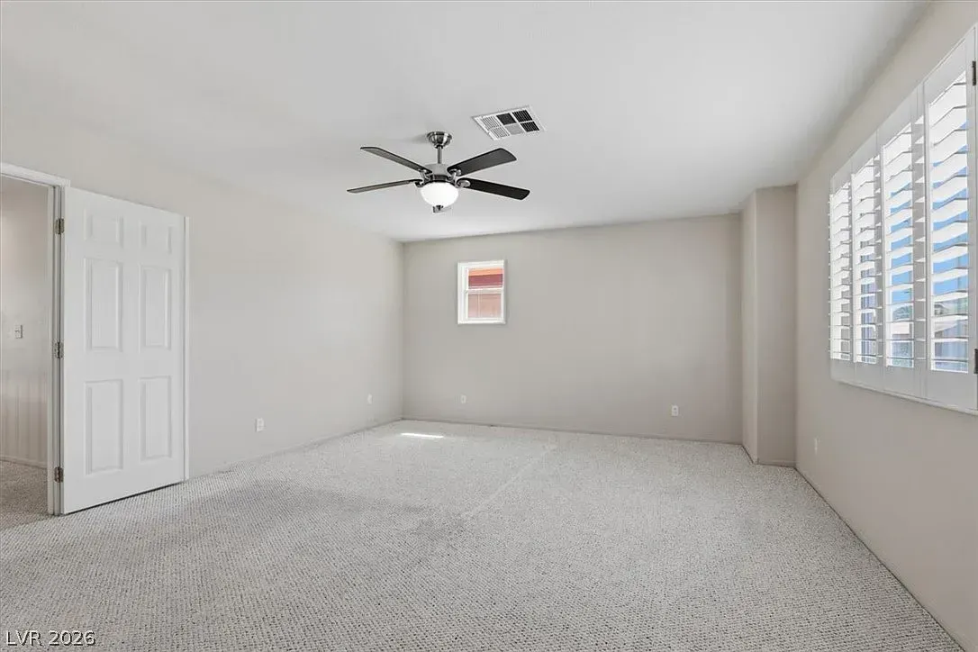 Empty beige bedroom with white carpet, ceiling fan, closet door, window shutters, and small wall art