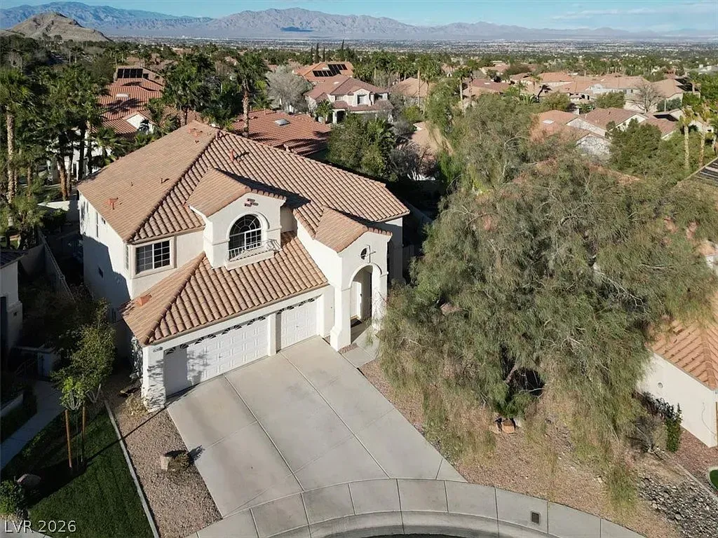 Suburban house with beige roof and driveway, surrounded by trees under a clear sky.