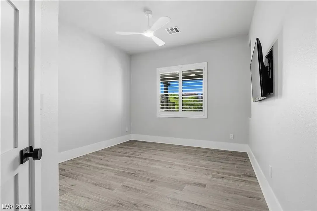 Empty white bedroom with gray wood floor, window, ceiling fan, and wall-mounted TV