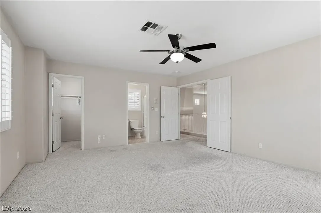 Empty beige bedroom with carpet, ceiling fan, and open closet and bathroom doors
