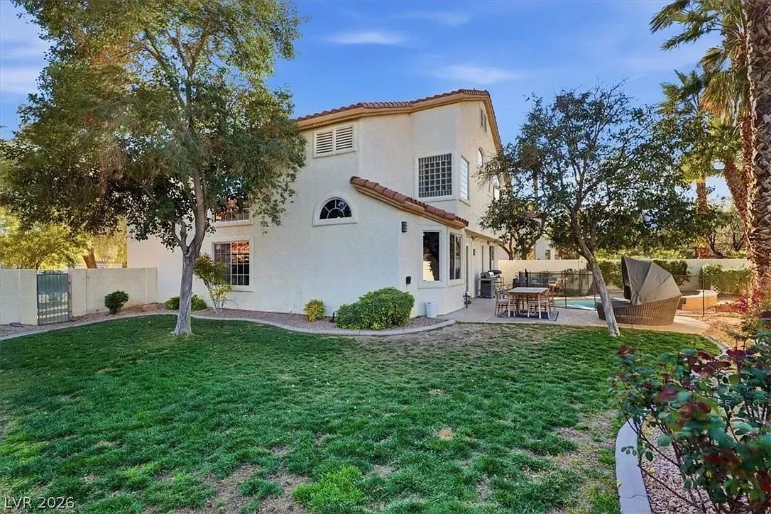 White two-story house with red-tiled roof, green lawn, and patio furniture under a sunny blue sky
