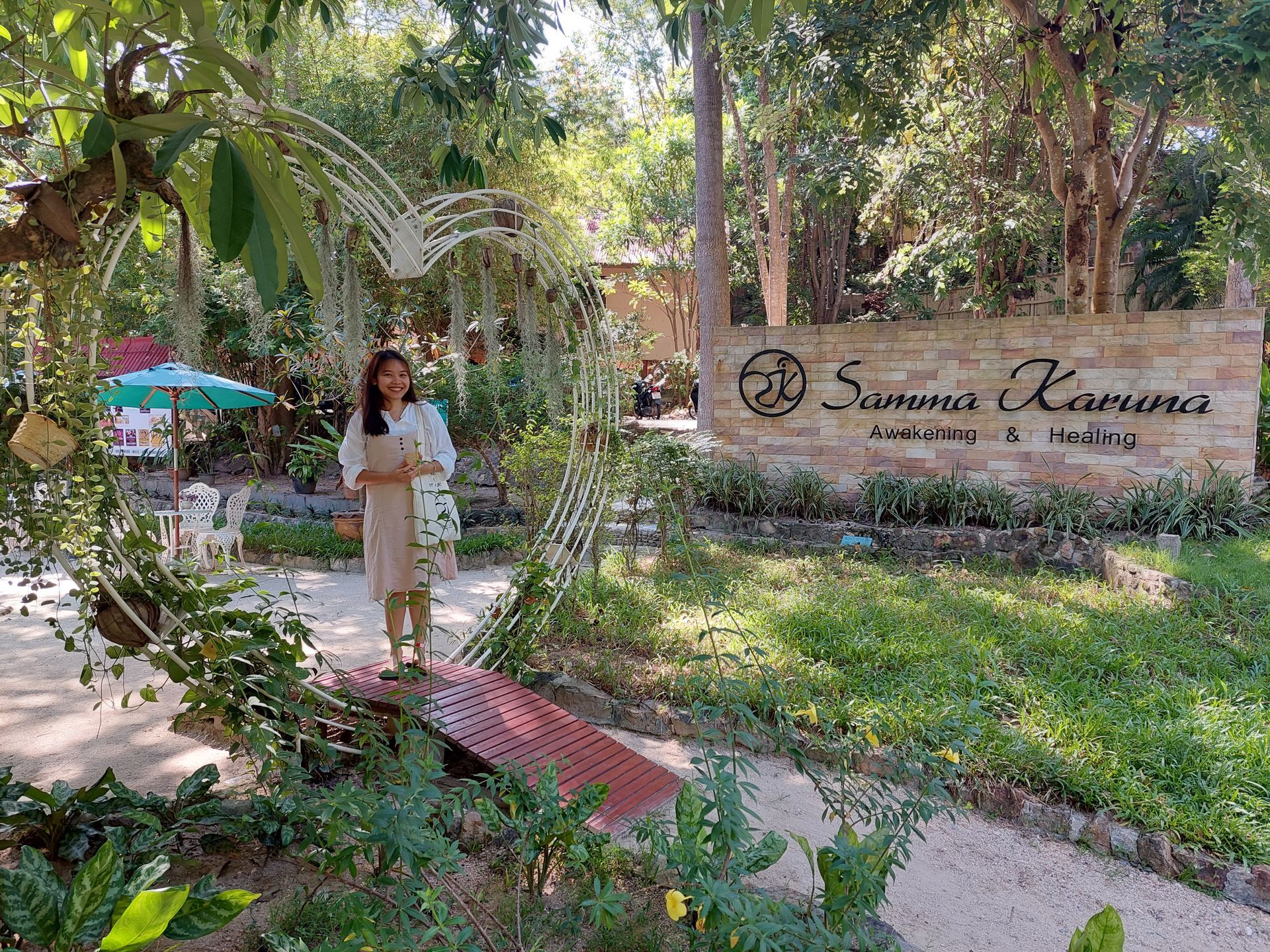 Woman walking along a garden path beside a sign reading “Swana Kamana” in a lush, tropical setting