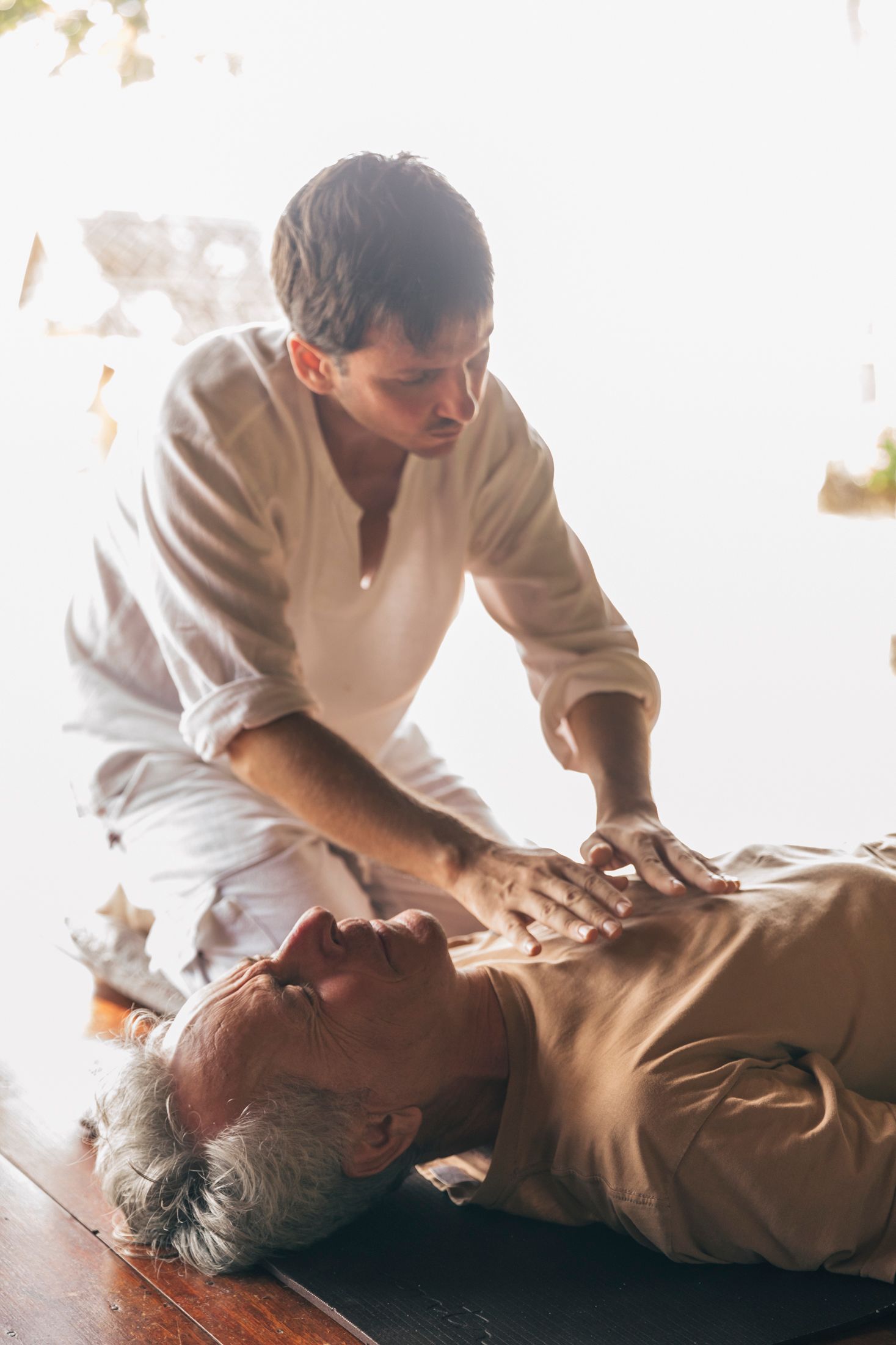 Person performing chest compressions on an older man lying on the floor in a bright room
