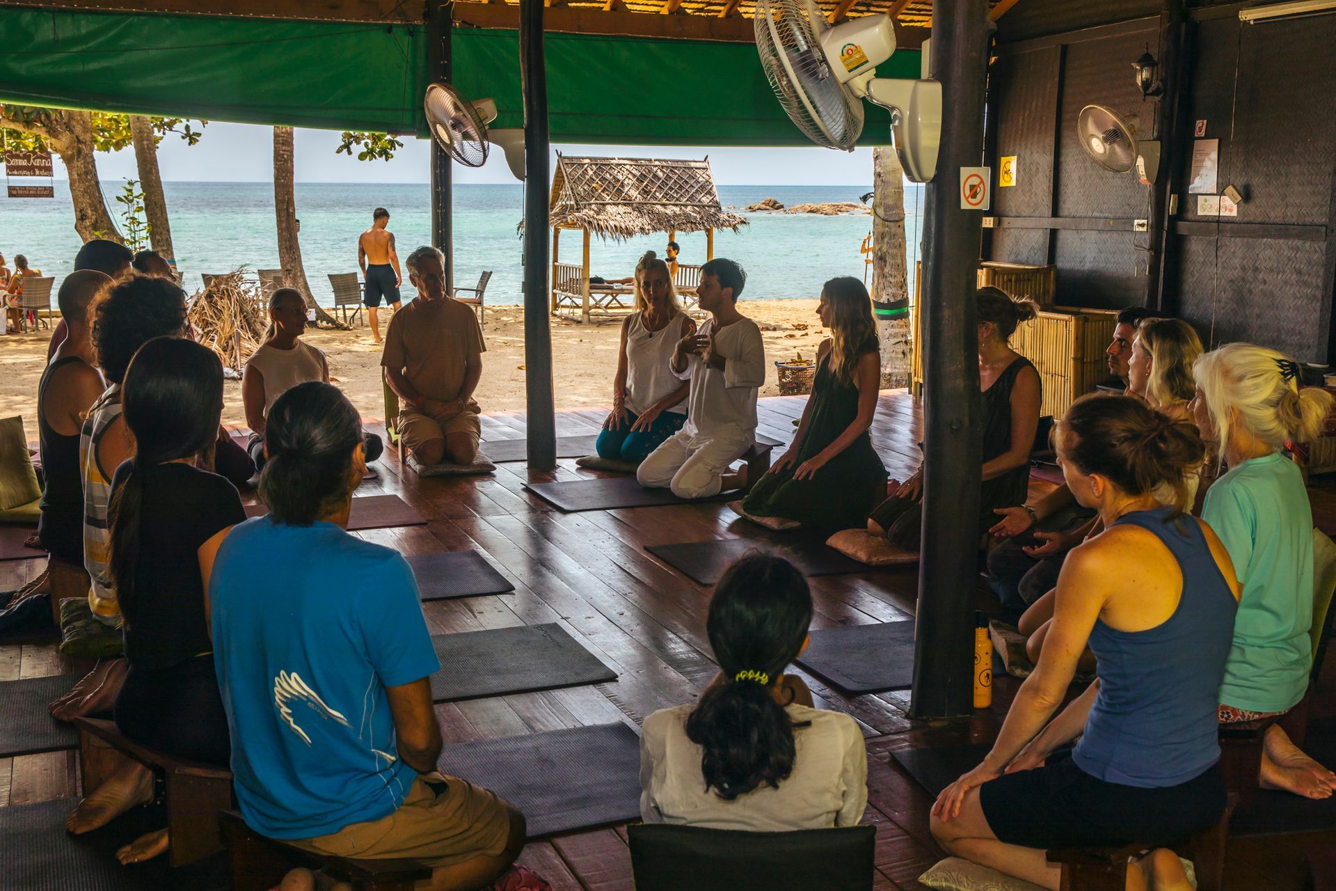 People seated in a beachside yoga class under a shaded pavilion, facing the ocean