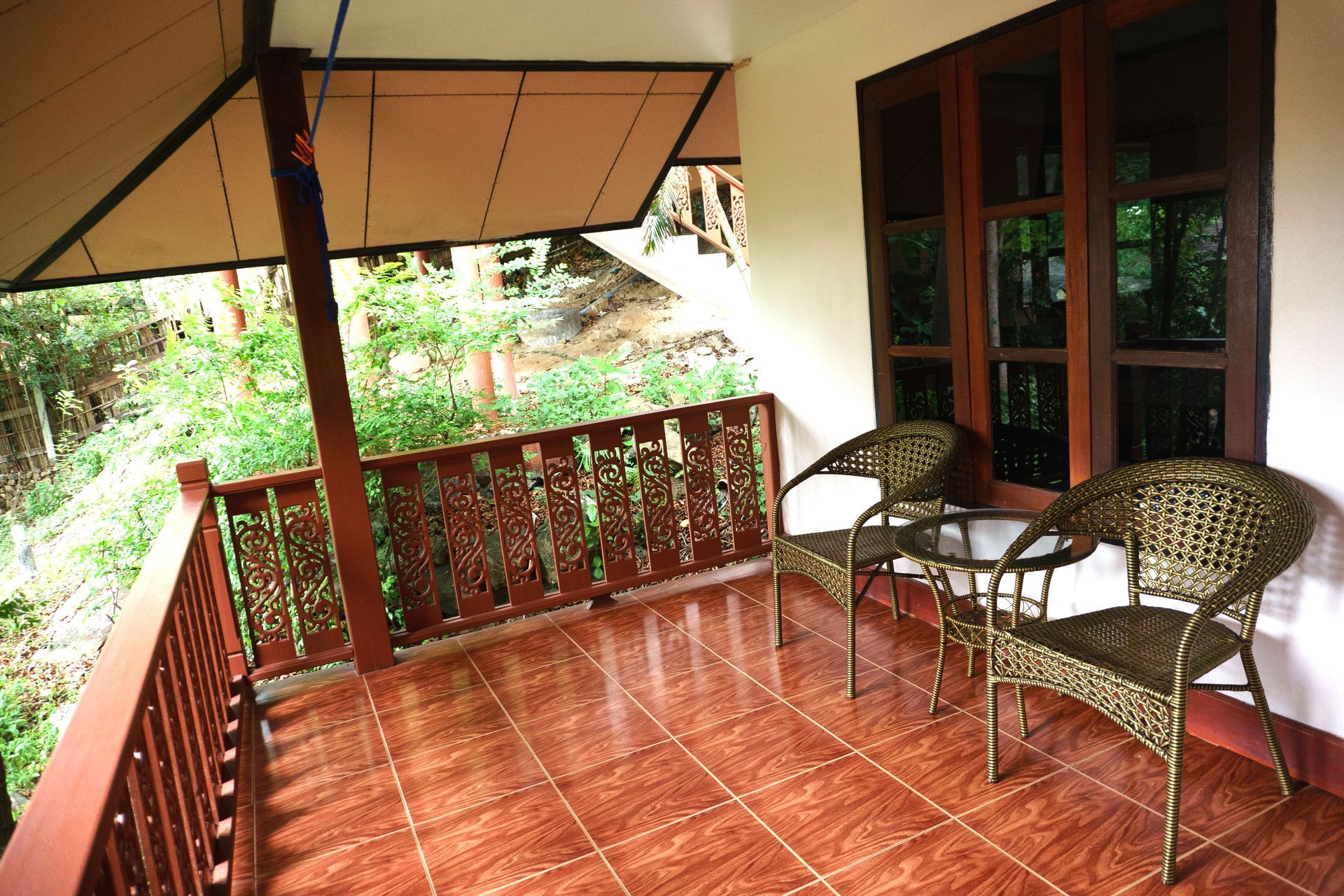 Covered porch with tiled floor, wicker chairs, a table, and a view of lush greenery beyond the railing