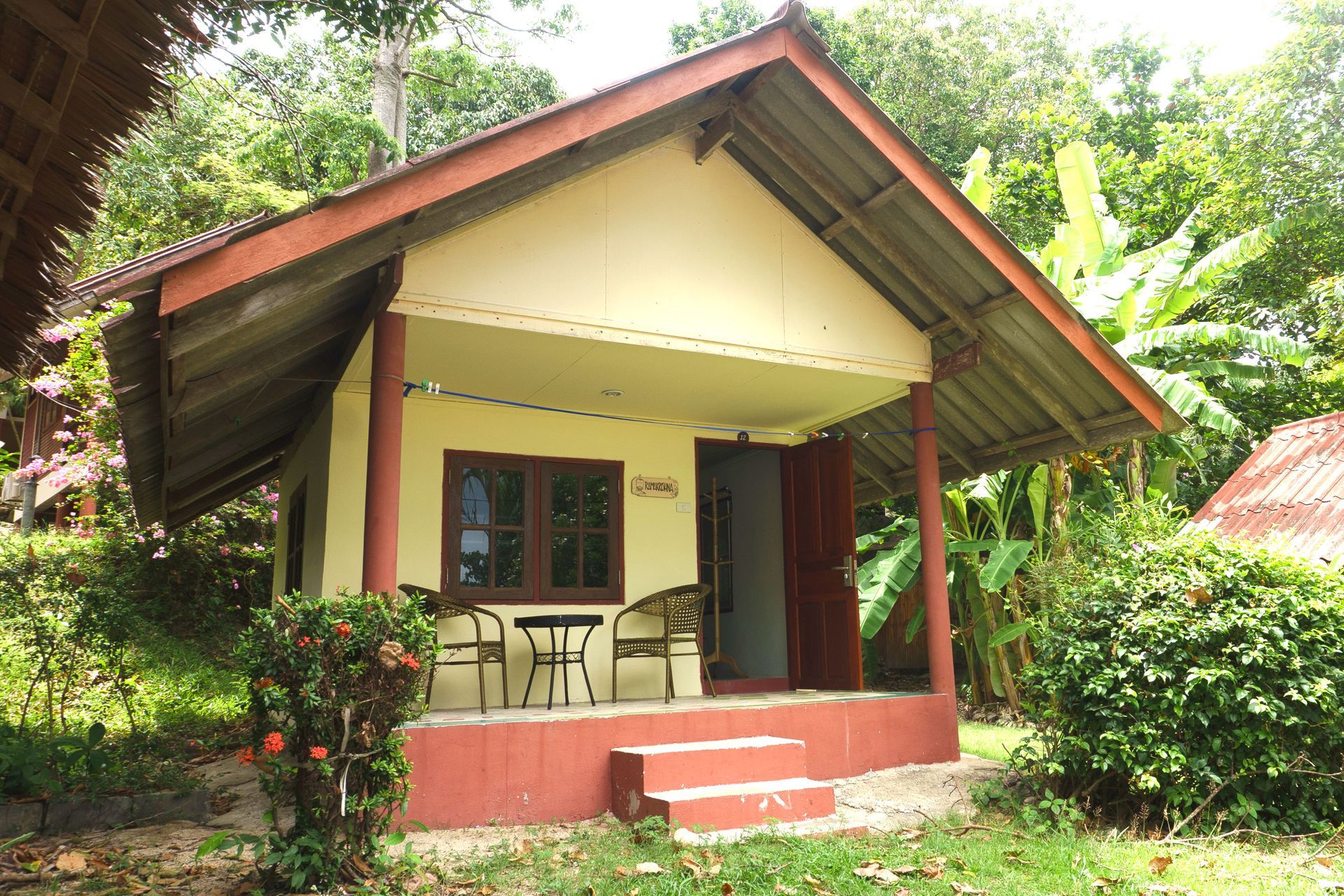 Small cream house with a red roof and porch, surrounded by lush green vegetation.