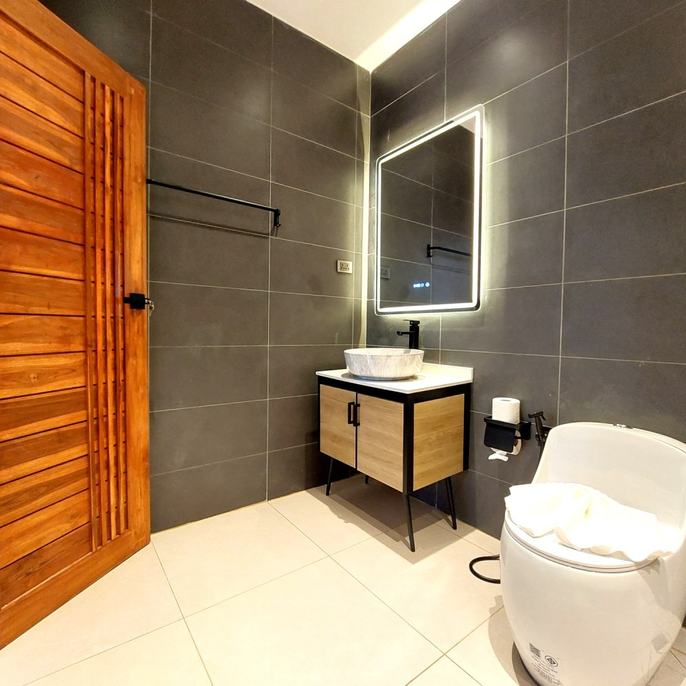 Modern bathroom with tiled walls, wood vanity, illuminated mirror, towel rack, and toilet paper holder
