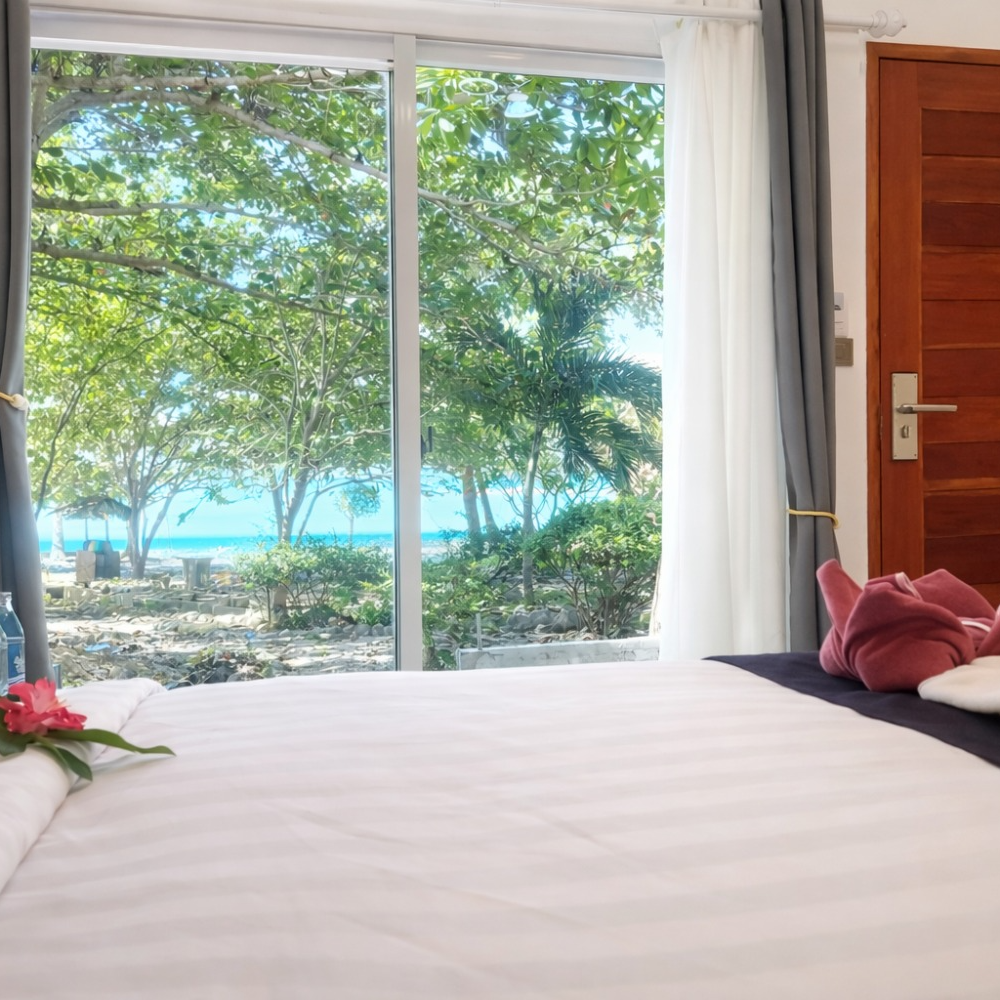 Bedroom with white bed, open window view of tropical trees and blue ocean, and a wooden door at right