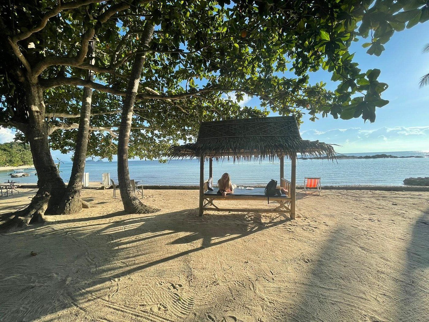 Shaded beach hut with two people sitting on a sandy shore by calm blue water at sunset