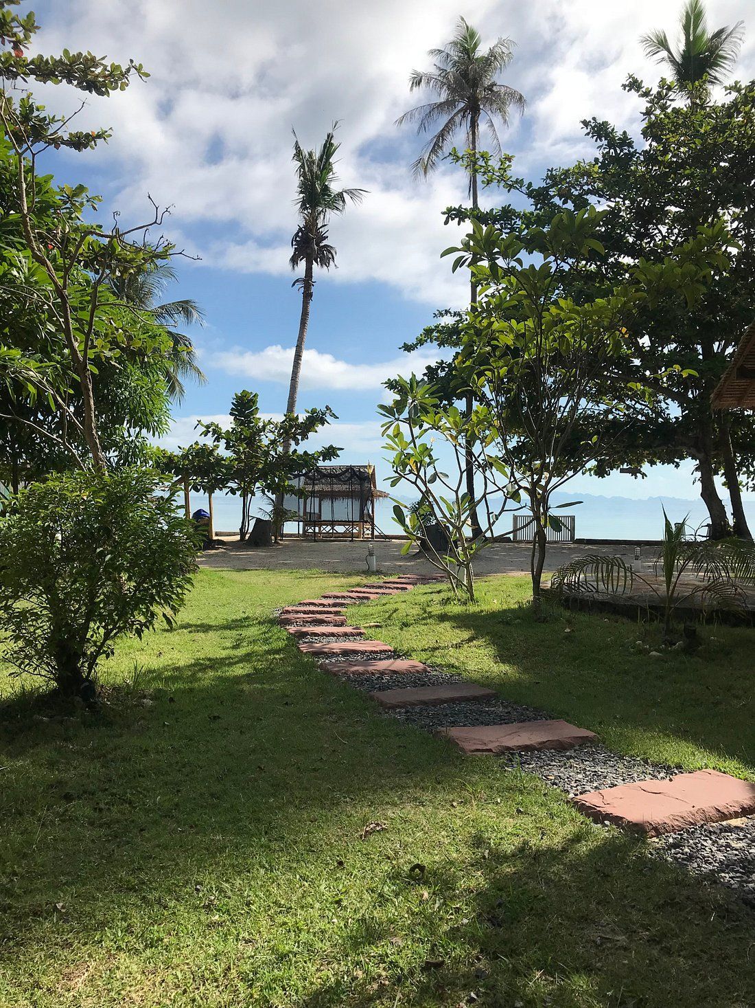 Tropical garden path leading to a small beachside pavilion under palm trees and blue sky