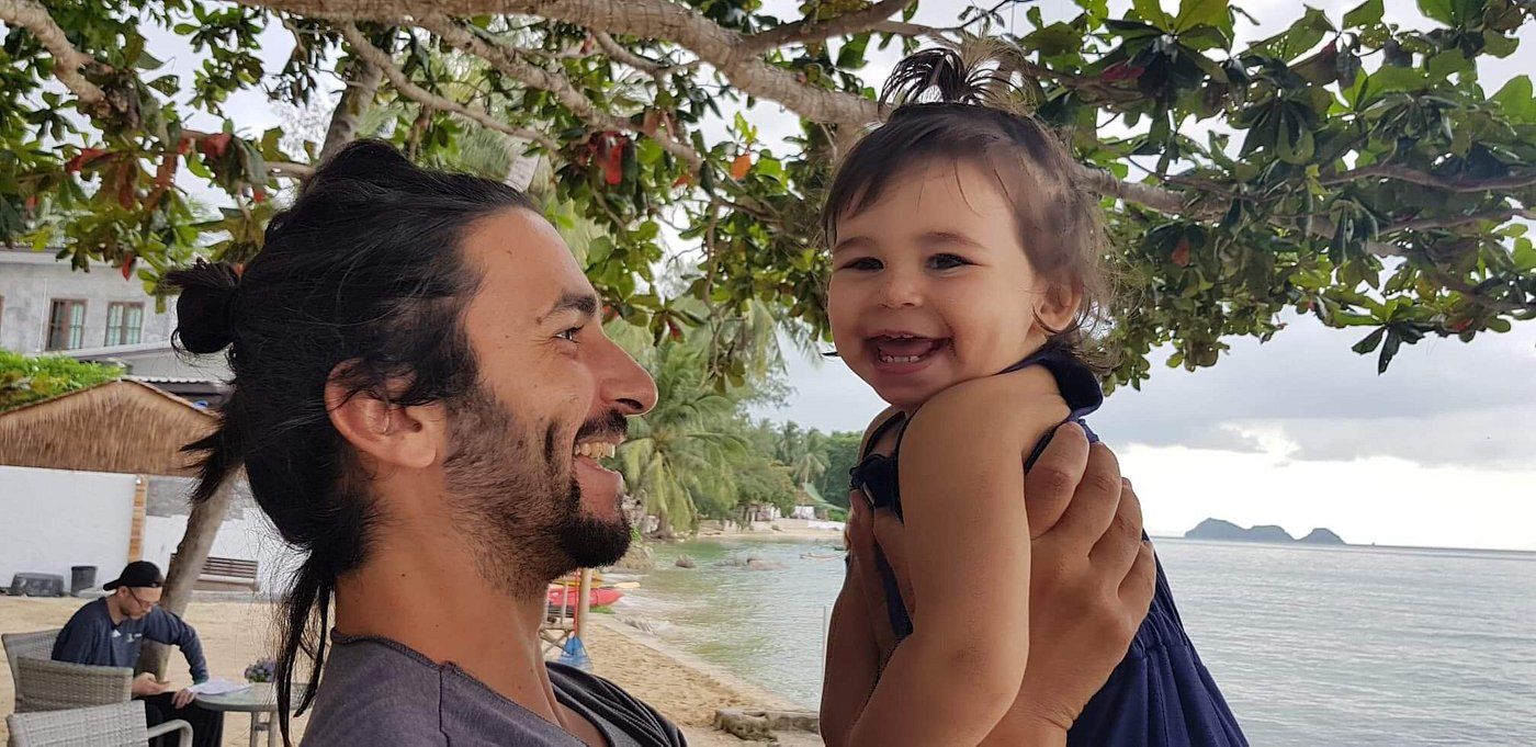 Man holding a smiling child outdoors under a tree, with water and distant mountains in the background