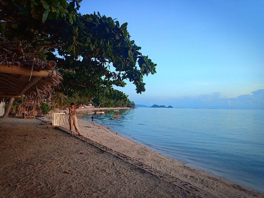 Quiet tropical beach with a thatched hut, overhanging trees, calm blue water, and distant islands at sunset